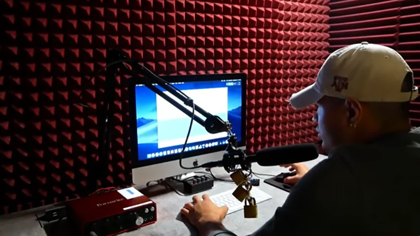 A person in a Texas A&M baseball cap records at a desk with a microphone and audio interface in a red soundproofed studio.